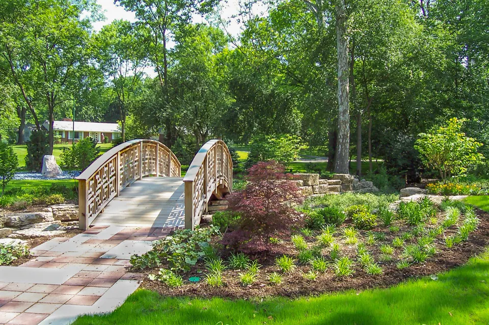 Paver garden walk leading toward a small arched wood bridge with low stone walls and layered planting